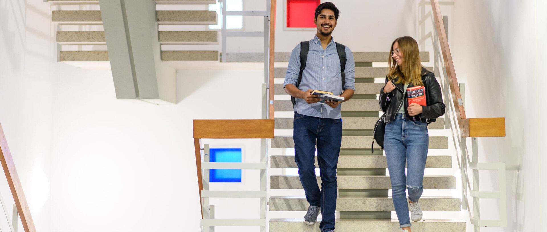Zwei Studierende gehen lächelnd eine Treppe herunter. Der linke Person blickt in die Kamera, trägt ein graues Hemd, dunkle Jeans und einen Rucksack, während sie einige Bücher hält. Die andere Person trägt eine dunkle Jacke, eine helle Jeans und hält einen Duden in der Hand.