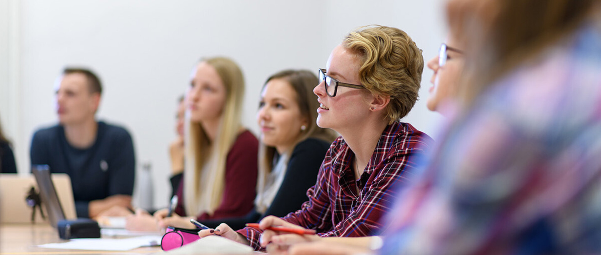 Studierende sitzen an einem Tisch und hören bei einem Vortrag zu.